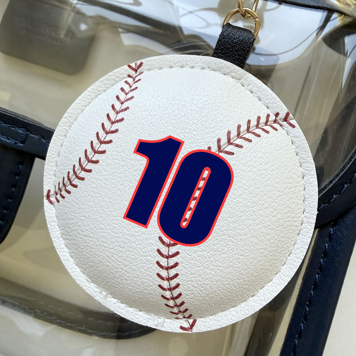 Baseball-shaped keychain, handbag accessory with red stitching on a faux-leather surface with customized vinyl lettering.