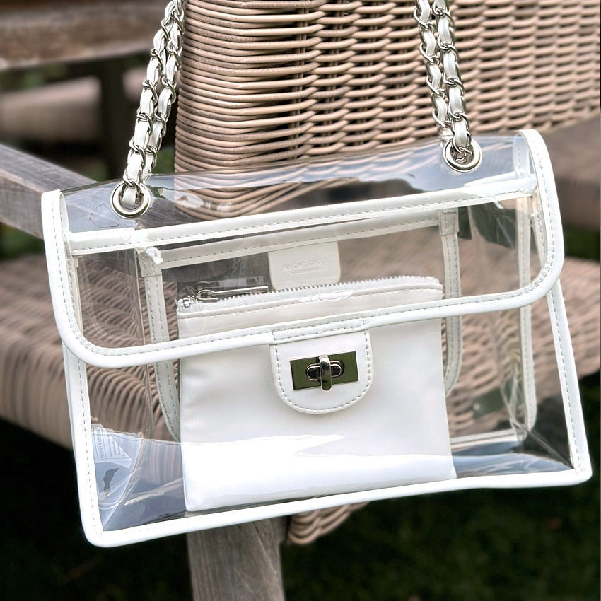 Clear, stadium-compliant handbag with white trim and silver chain strap hanging from a wicker lawn chair