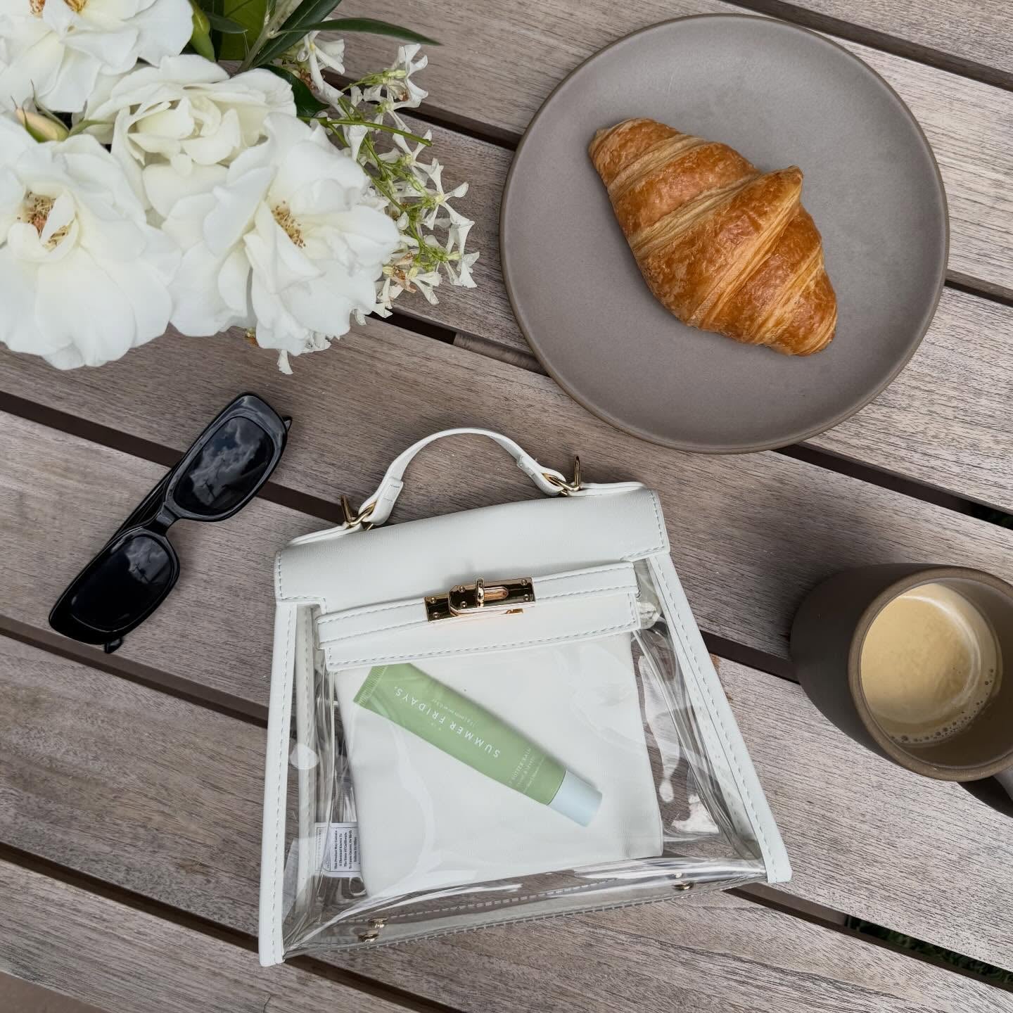 Clear stadium-compliant handbag with skincare product, croissant, sunglasses, and flowers on a wooden table to show purse size