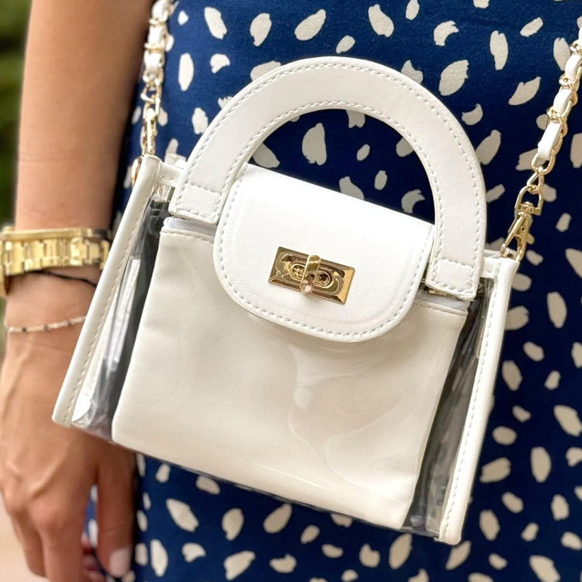 See though clear handbag with white trim and gold hardware being worn by a woman in a blue dress with white polkadots.