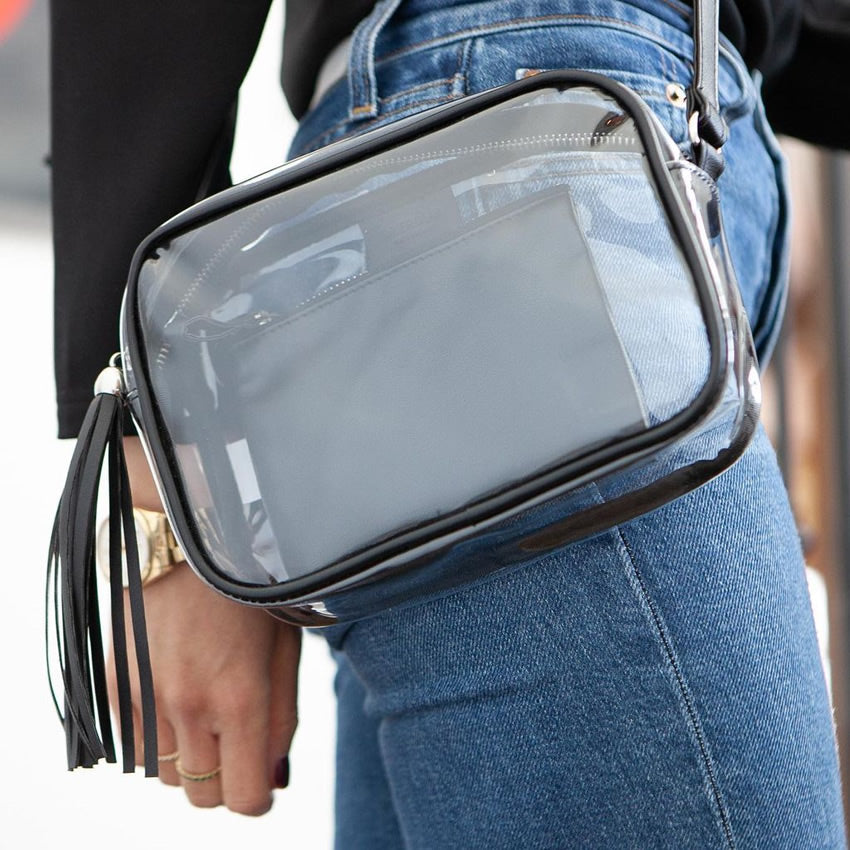 Clear crossbody purse with black tassel held by a person wearing blue jeans.