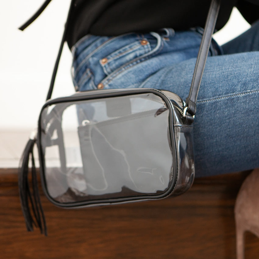 Clear handbag with black strap worn by a person sitting on a wooden floor.