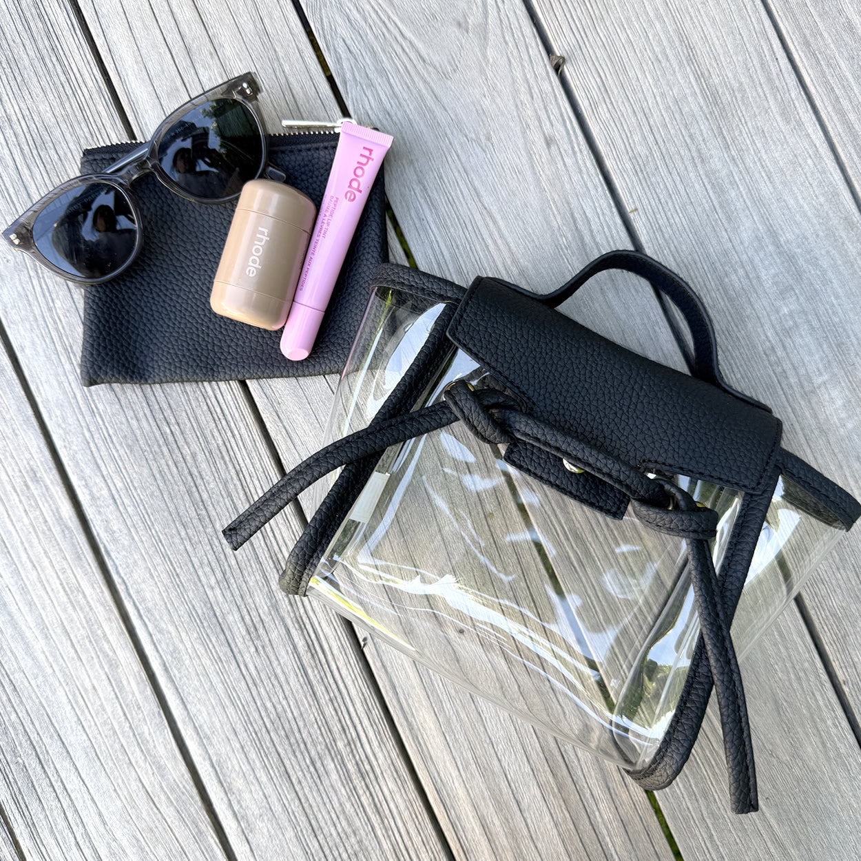 Clear, stadium approved bag with black straps, sunglasses, and lip products on a wooden surface