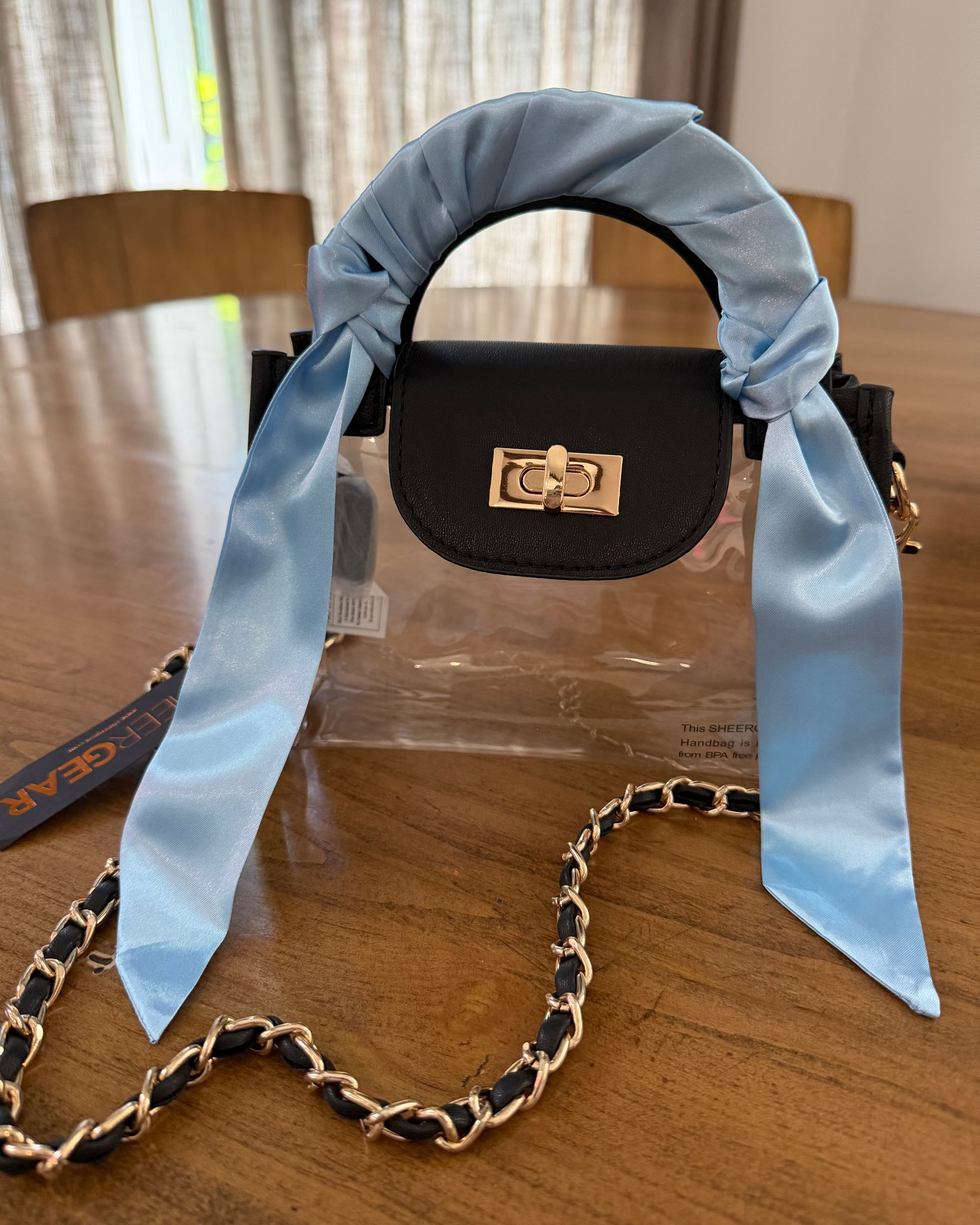 A black leather handbag with a light blue satin handle wrap and matching scarf, featuring a gold turn-lock clasp and a black-and-gold chain strap, displayed on a wooden table.