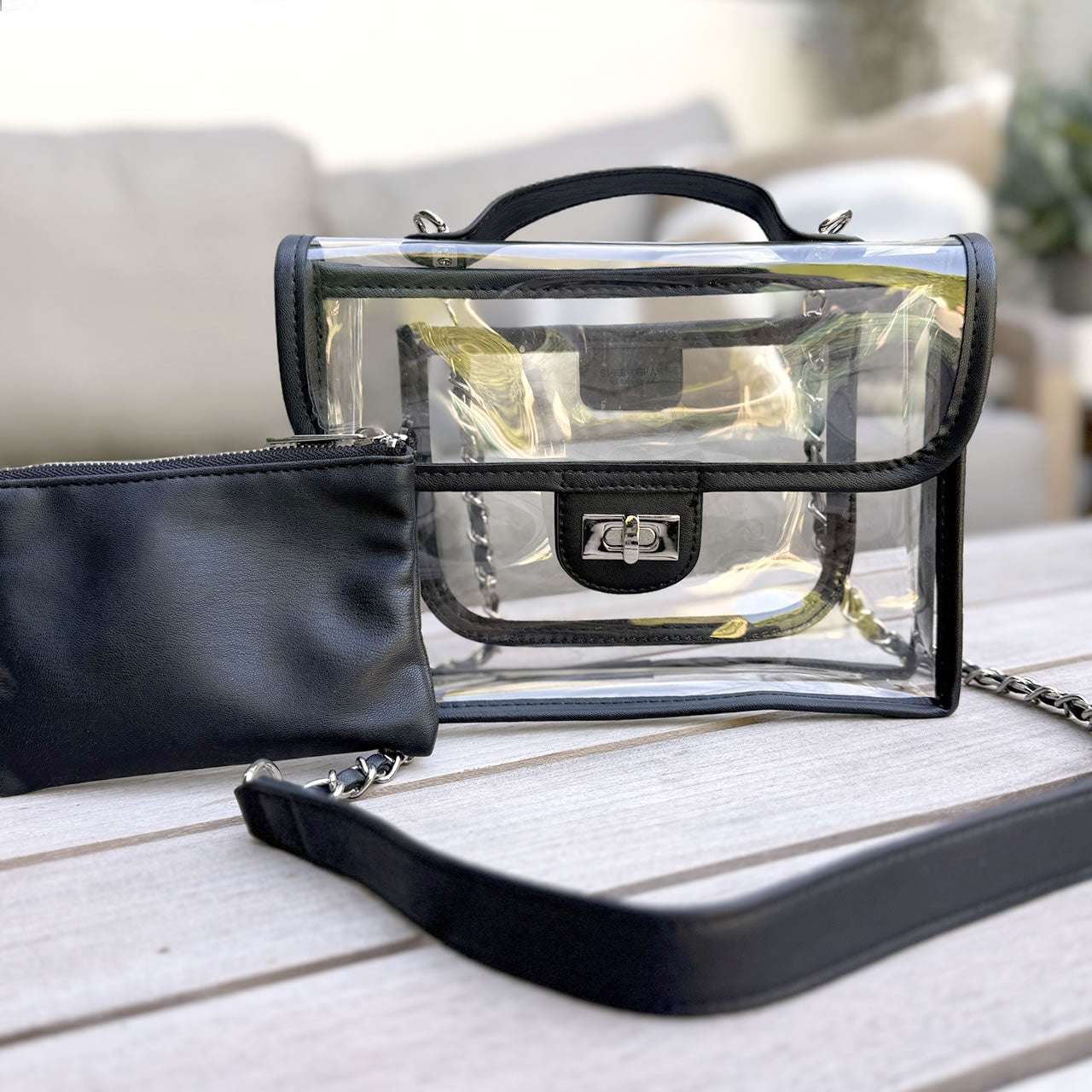 Black handbag with clear section and chain detail on a wooden surface with plants in the background
