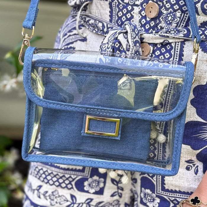 Blue denim, see-through purse with gold accents worn by a person in a blue patterned outfit.