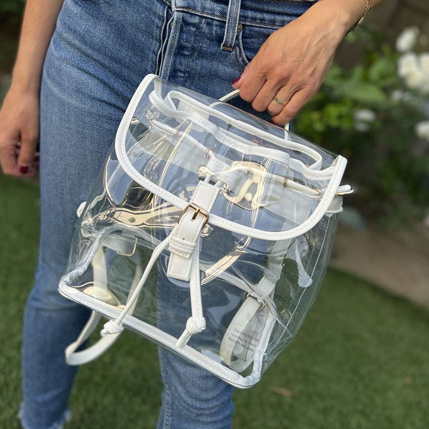 Clear backpack with white straps held by a person wearing blue jeans.