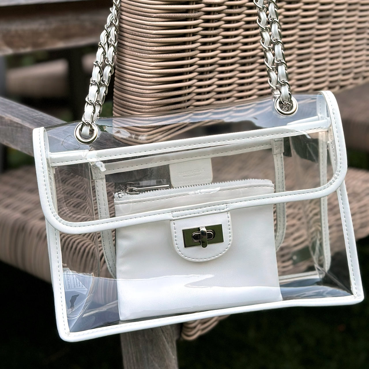 Clear, stadium-compliant handbag with white trim and silver chain strap hanging from a wicker lawn chair