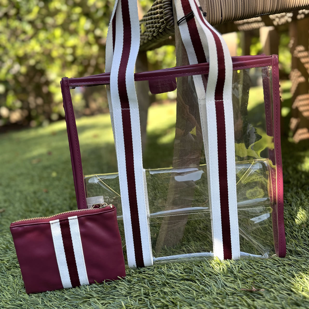 Clear stadium tote bag with maroon and white striped handles and a matching small pouch on grass.