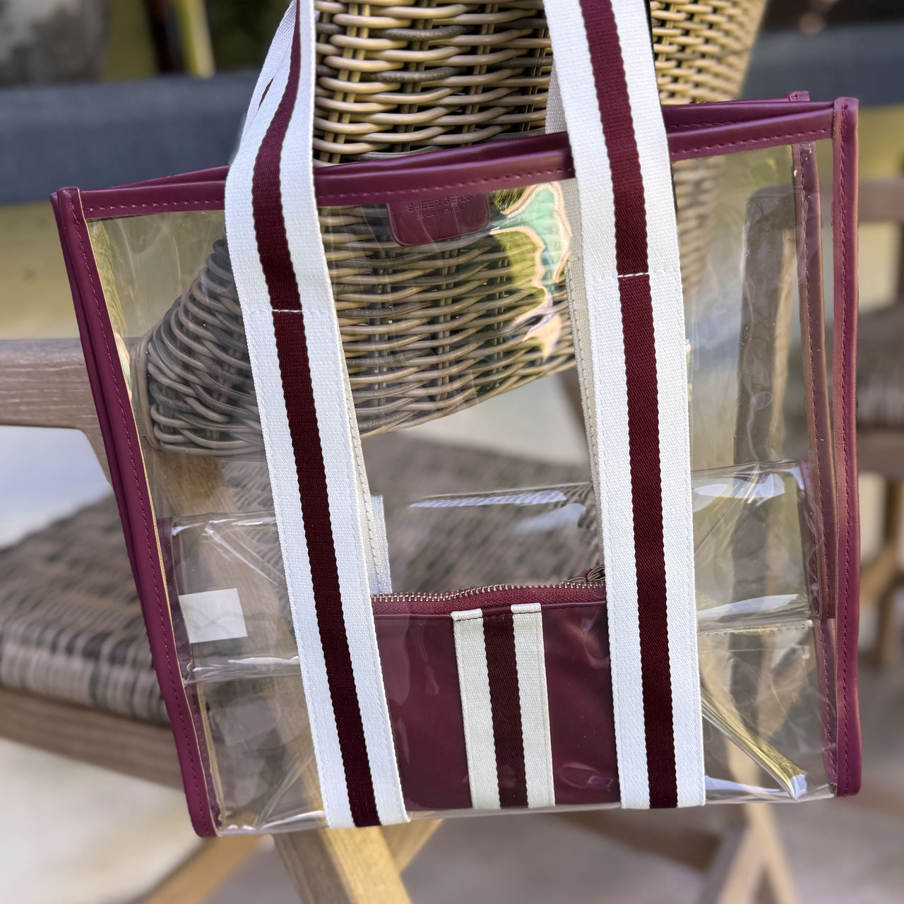 Clear stadium approved tote bag with maroon straps over a wicker chair
