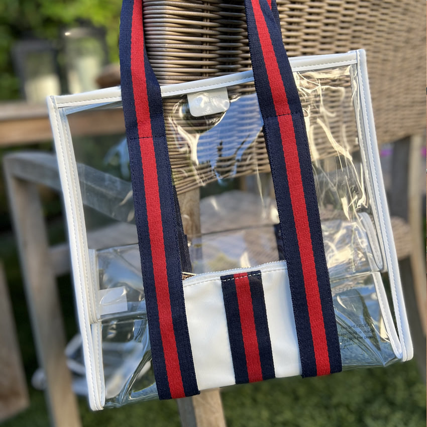 Clear, stadium-compliant tote bag with red and blue striped strap on a blurred outdoor background