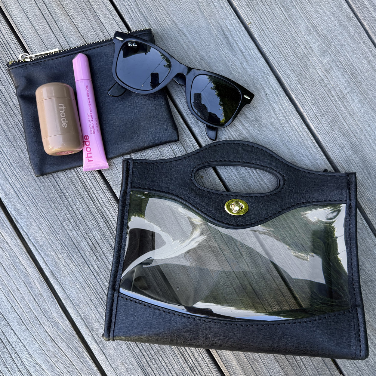Black, clear, stadium approved purse with sunglasses and makeup items on a wooden surface