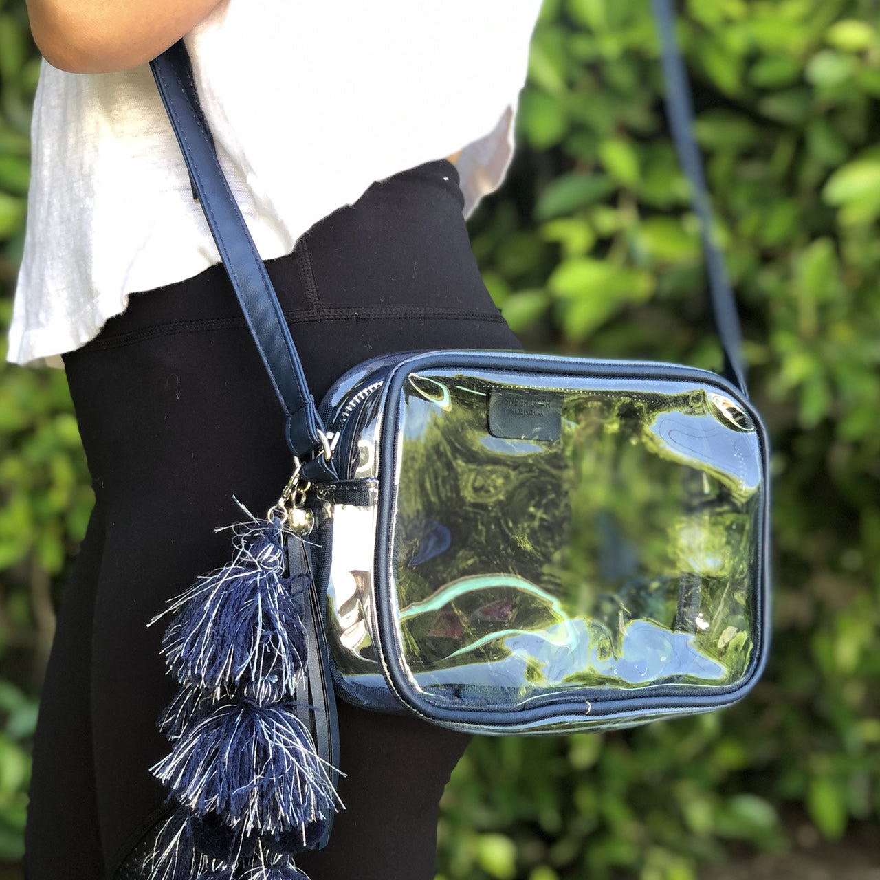 Clear, stadium approved bag with navy trim and tassle worn crossbody by a woman in yoga pants