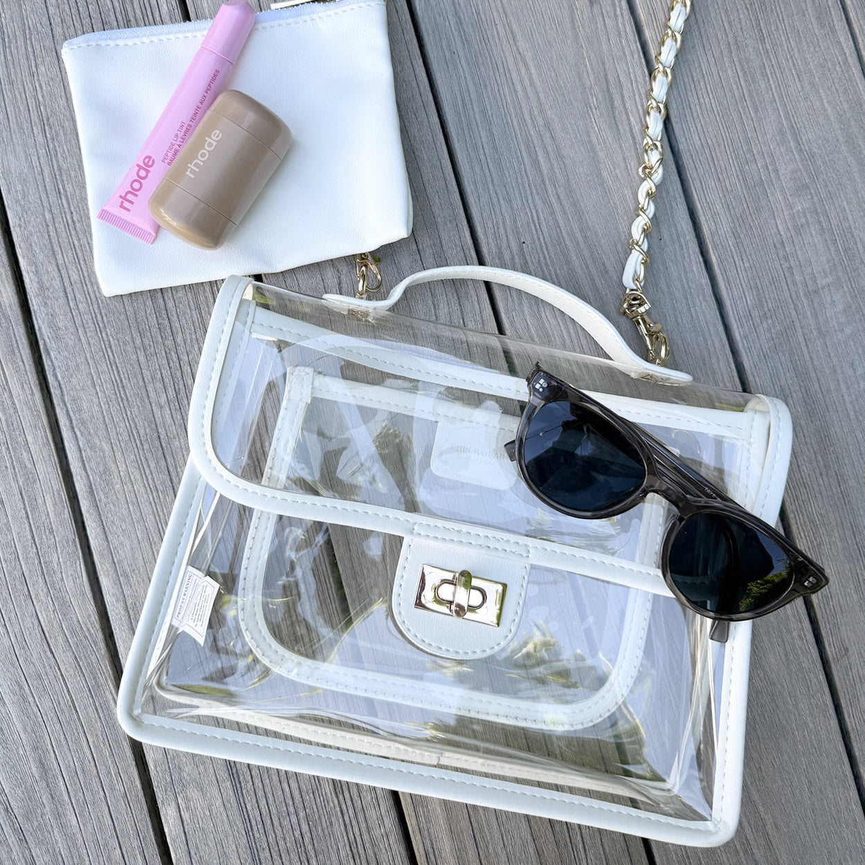Clear stadium-approved purse flat lay with sunglasses and lipstick on a wooden surface