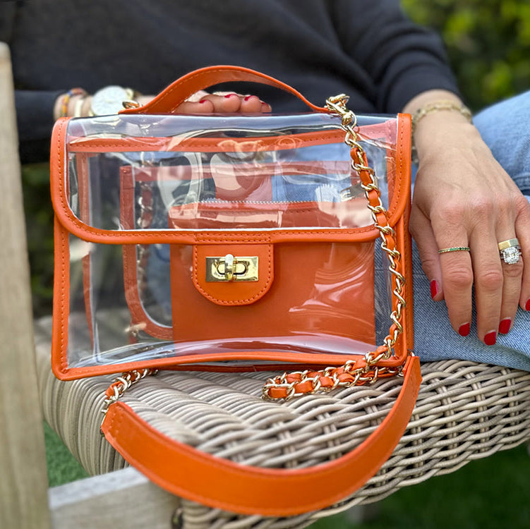 A person holding a stadium-approved, orange-trim crossbody bag with gold-colored accents and a chain strap.